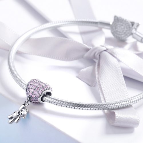 Elegáns fehér masnis ajándékdoboz tetején pihenő ezüst kígyólánc karkötő, rajta egy csillogó rózsaszín szíves és játékmacis függő charm díszítéssel.