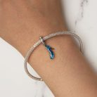 Közeli felvétel egy női csuklón viselt hálós ezüst karkötőről, amelyen egy elegáns, kék színű magassarkú cipő függő charm látható fehér márvány háttér előtt.