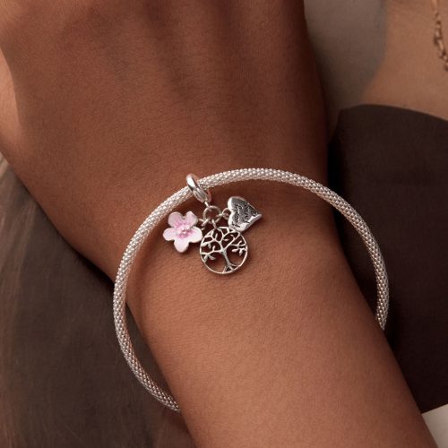 Közeli felvétel egy női kézen lévő ezüst karkötőről, amelyen a részletgazdag, rózsaszín virágos és „Family forever” feliratú szívvel díszített életfa charm látható.