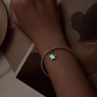 Női csuklón lévő ezüst karkötő, amelyen a csillag alakú függő charm intenzív zöld fénnyel világít a sötétben.