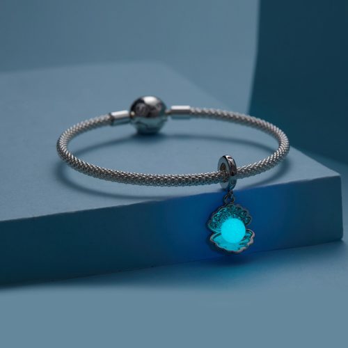 Ezüst karkötőre helyezett gyöngykagyló charm, amelynek központi gyöngye élénk kékeszöld fénnyel világít a sötétben.
