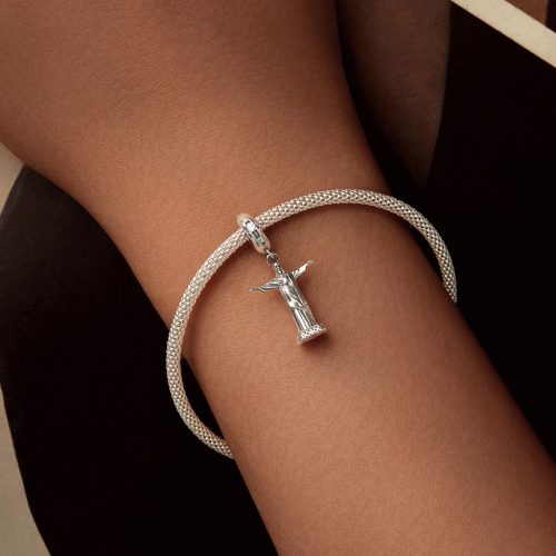 Női csuklón viselt hálós ezüst karkötő a riói Megváltó Krisztus szobor függő charmmal, elegáns, sötét tónusú háttér előtt.