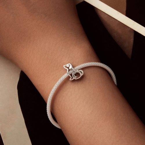 Női csuklón viselt hálós ezüst karkötő az absztrakt kutyás sziluett charmmal, elegáns, sötét tónusú környezetben.